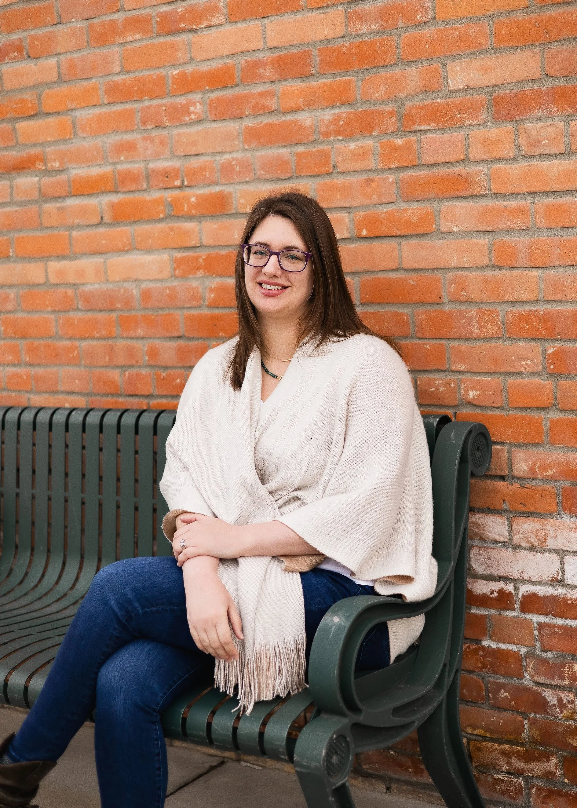 A woman sitting on a green park bench in front of a red brick wall, smiling, wearing glasses, a cream cardigan, and blue jeans.