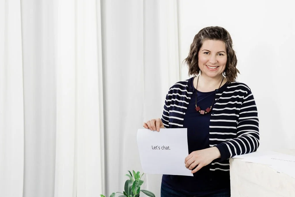 A woman smiling and holding a sign that says "Let's chat" in front of a white curtain background.