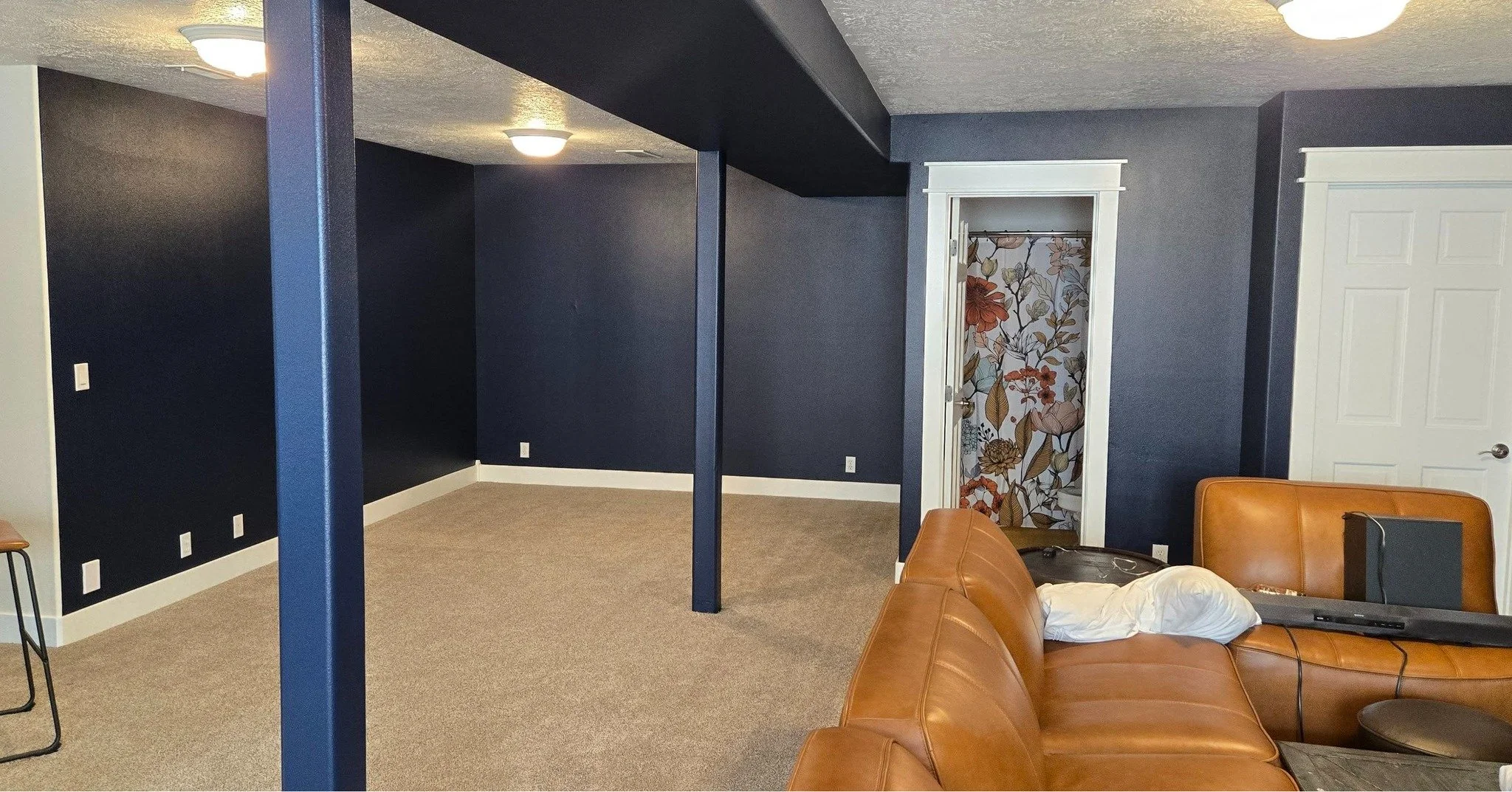 A living room with navy blue walls, beige carpet, white trim, and a doorway with floral-patterned curtains. There is a brown leather sofa with a white pillow, a laptop, and an orange chair. The ceiling has round white light fixtures.