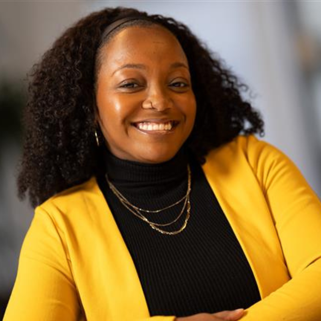 A woman with curly hair smiling, wearing a yellow blazer over a black turtleneck, accessorized with layered gold necklaces and earrings.