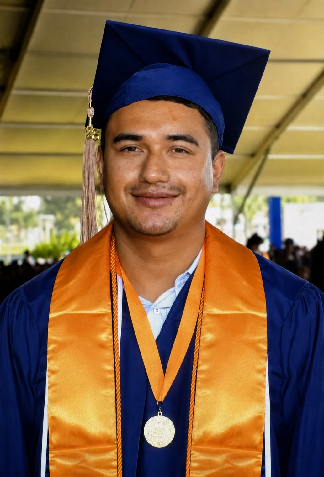 A man in a blue graduation cap and gown with an orange stole and medal at a graduation ceremony.