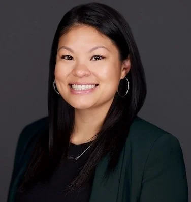 A woman with long black hair, wearing a dark blazer and silver hoop earrings, smiling against a dark gray background.
