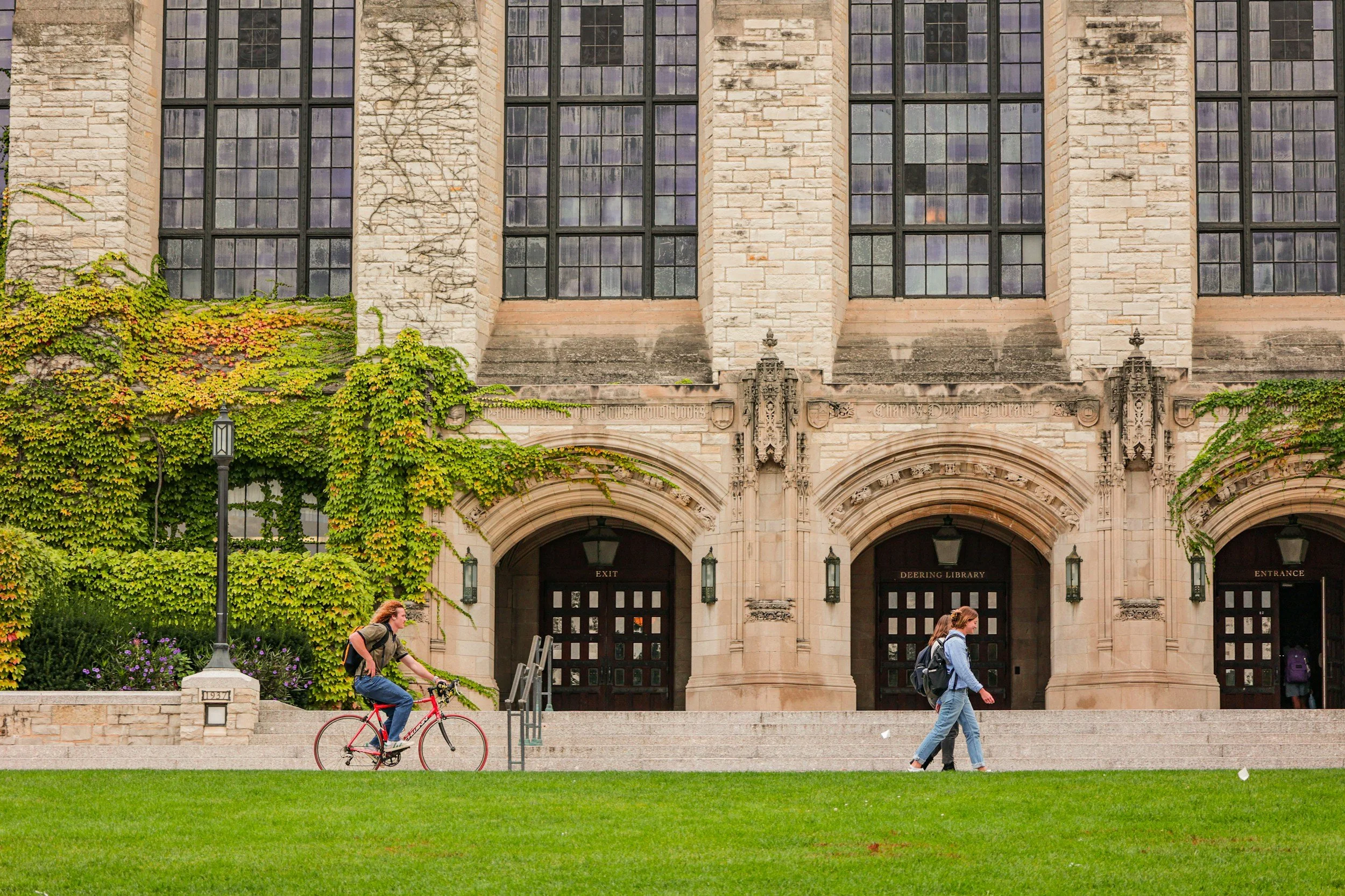 People walking and riding a bike in front of a historic stone library building with large windows, ivy, and decorative architectural details.