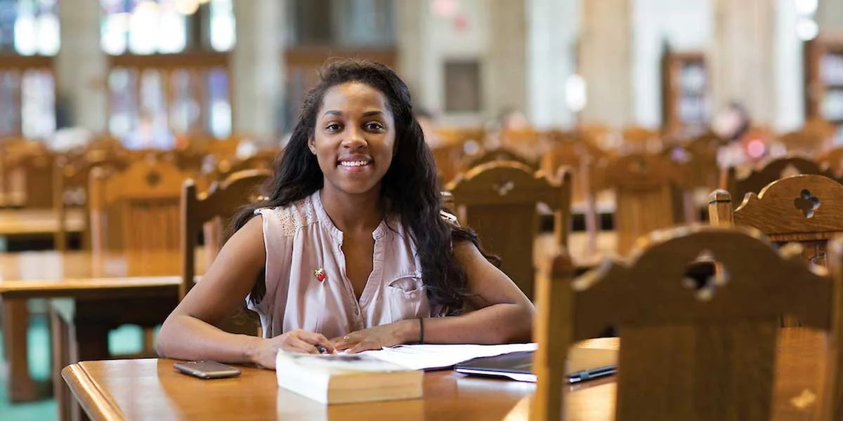 A young woman sitting at a wooden table in a library, smiling, with books and a notebook in front of her.
