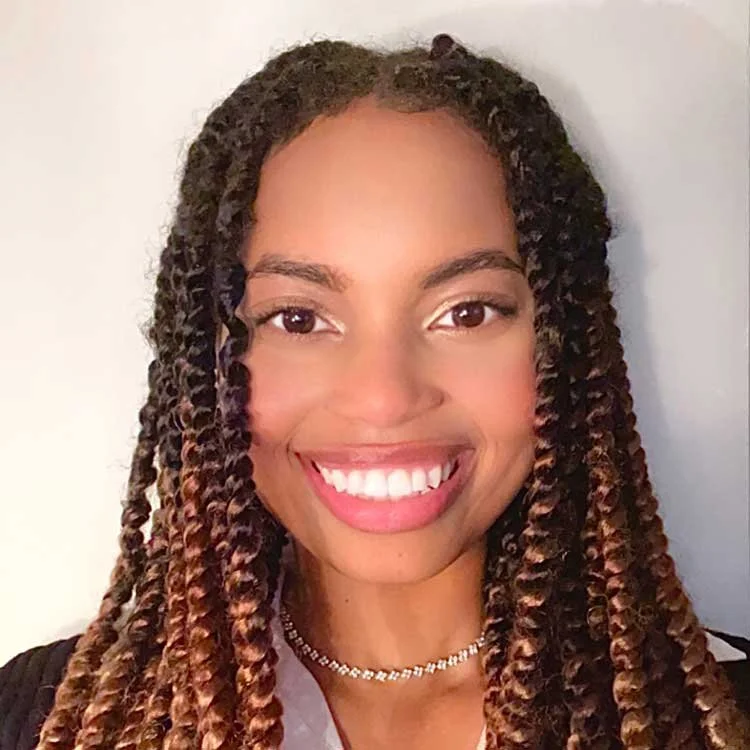 A young woman with long braided hair, smiling with white teeth, wearing a necklace and a black top.