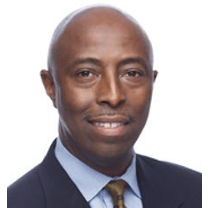 Portrait of a bald man wearing a dark suit, light blue shirt, and patterned tie, smiling against a white background.