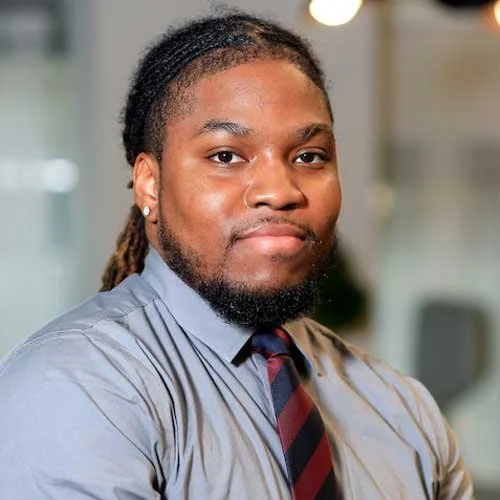 Professional portrait of a young African American man with dreadlocks, wearing a gray dress shirt and a striped tie, smiling confidently in an office setting.