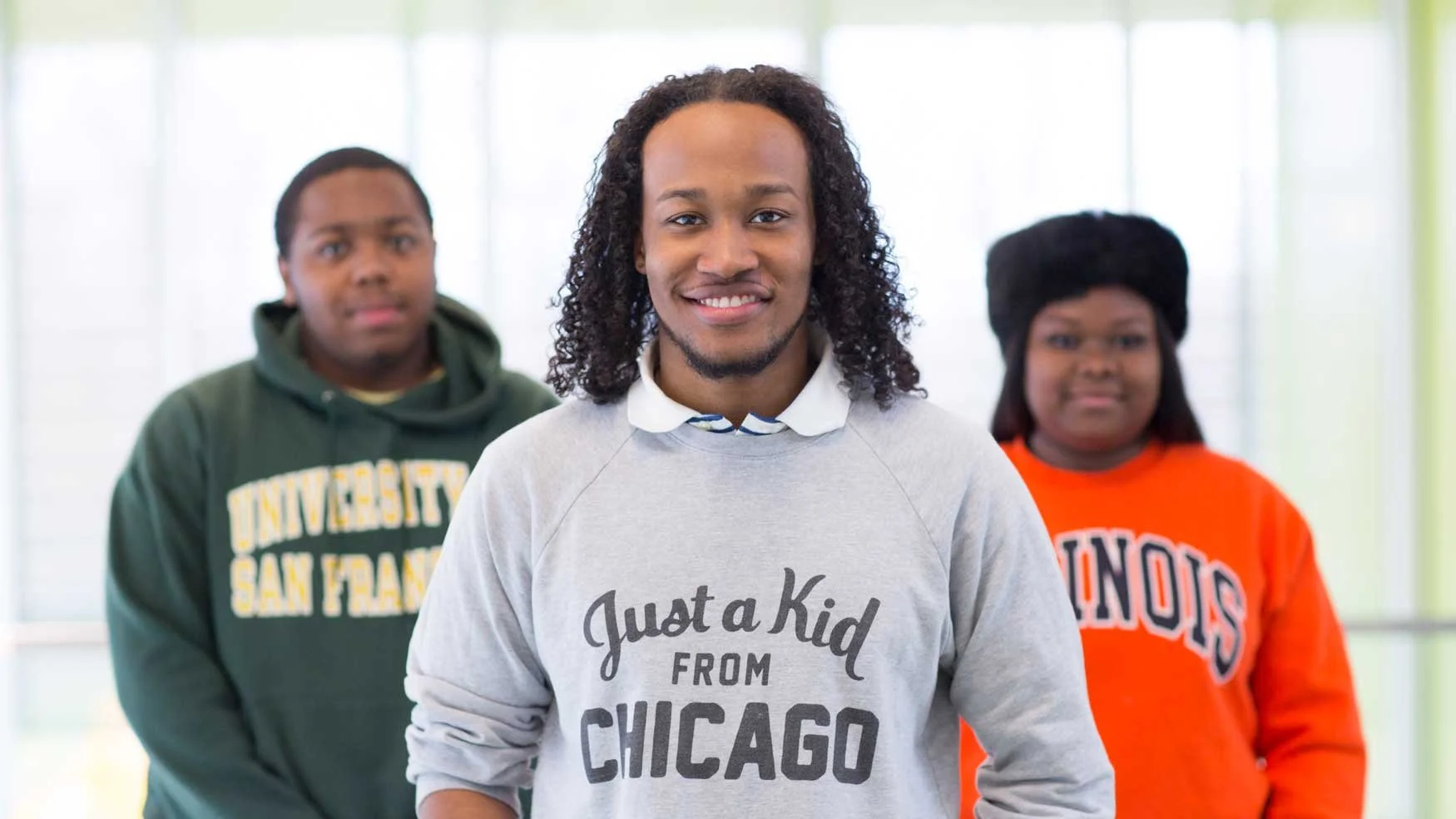 Three young people standing indoors with a bright background, smiling at the camera. The person in front wears a gray sweatshirt that says 'Just a Kid from Chicago.' Behind are a young man in a green hoodie with 'University of San Francisco' and a young woman in an orange sweatshirt with 'Illinois.'