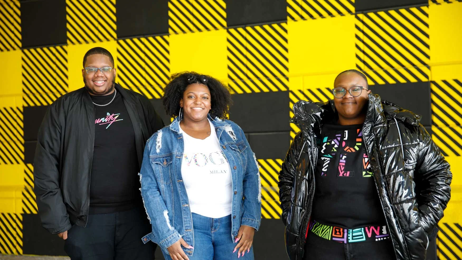 Three smiling people standing in front of a black and yellow patterned wall. The person on the left is a man wearing glasses, a black jacket, and a black shirt with pink and white text. The person in the middle is a woman with large curly hair, wearing a white shirt and a distressed denim jacket. The person on the right is a woman with short hair, wearing glasses and a shiny black puffer jacket with a desert-themed graphic shirt underneath.