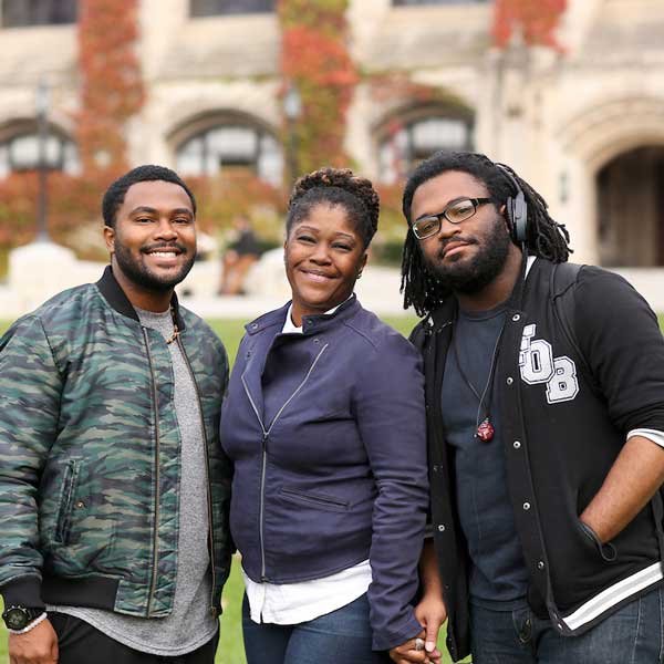 Three young adults standing outdoors in front of a historic building with red brick and arched windows. They are smiling and looking at the camera, dressed casually. The woman in the middle is wearing a navy jacket, the man on the left is in a camouflage jacket, and the man on the right is wearing headphones and glasses with a black jacket.