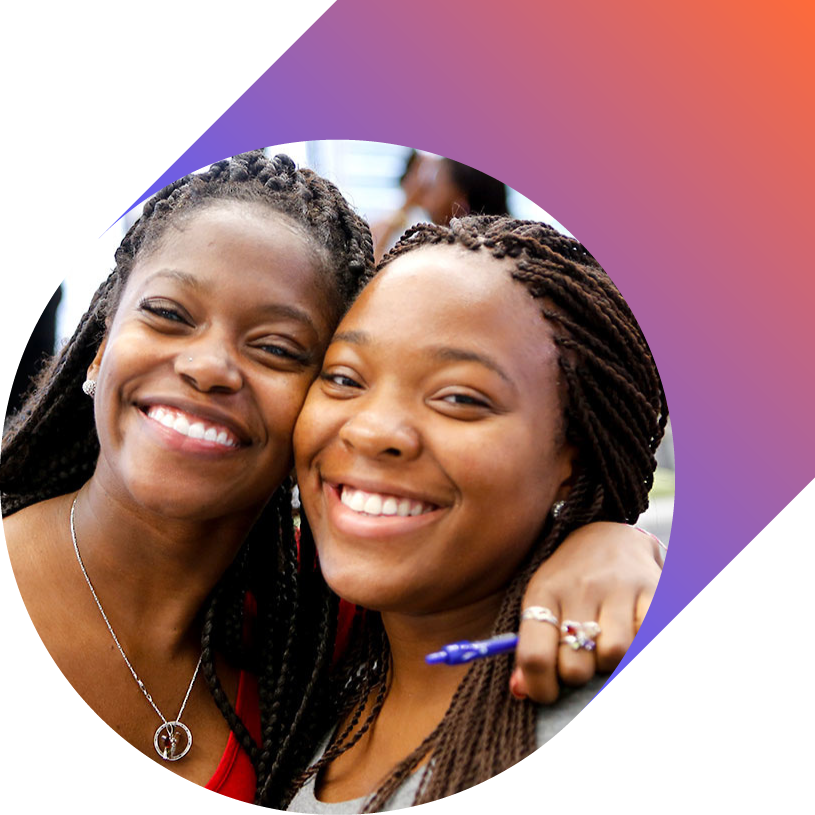 Two smiling women with braided hair embracing each other at an outdoor event.