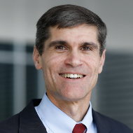 A smiling middle-aged man with dark hair, wearing a suit and tie, in an office setting.