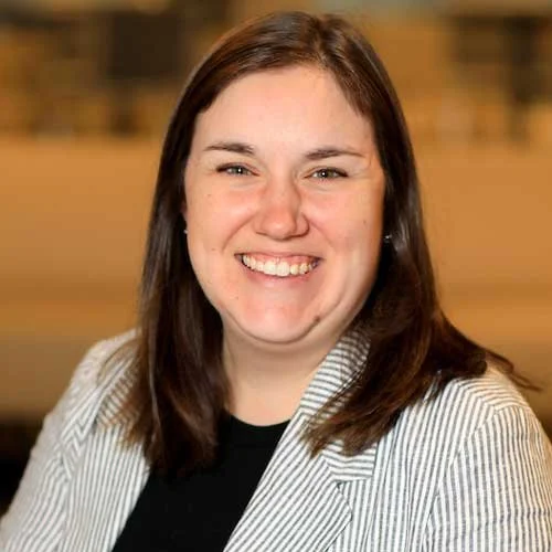 A woman with shoulder-length brown hair smiling, wearing a light-colored blazer and a black top, with a blurred indoor background.