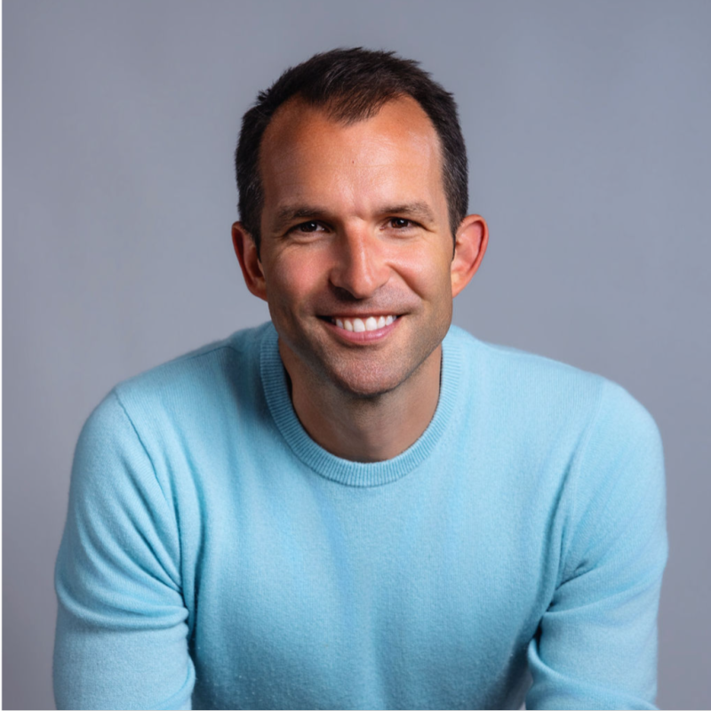 A smiling man with short dark hair, wearing a blue checkered shirt, against a dark blue background.