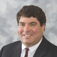 Professional headshot of a man in a suit smiling against a gray background.