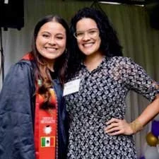 Two women smiling and posing together indoors, one wearing a red and black scarf, the other in a patterned dress.