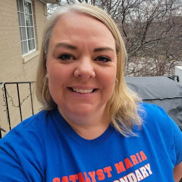 A woman with blonde hair taking a selfie outdoors on a balcony, wearing a blue T-shirt with red and white text.
