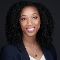 A woman with curly hair smiling, wearing a navy blazer and a necklace.