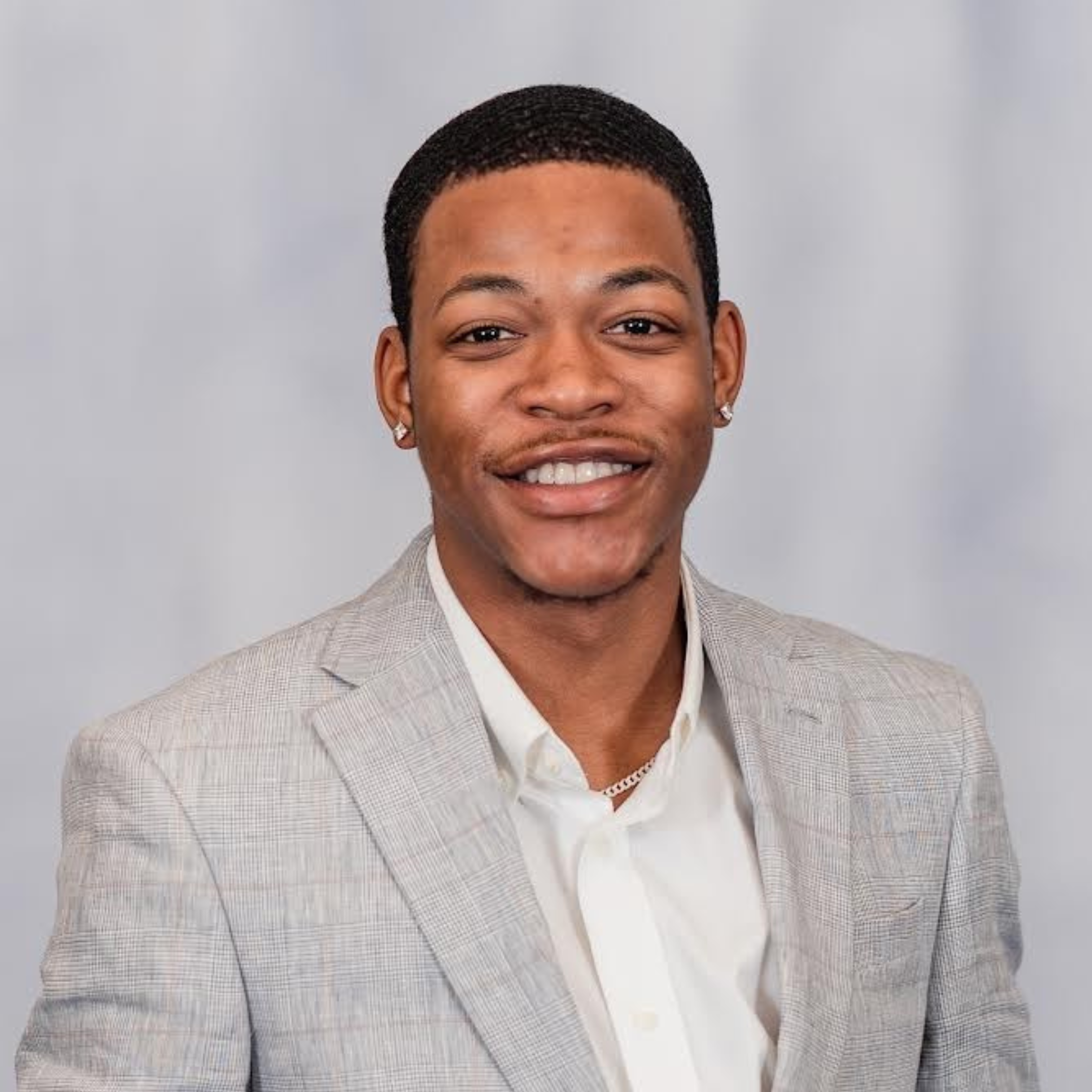 Close-up of a smiling African American man with short curly hair, wearing diamond earrings and a light-colored checkered blazer.