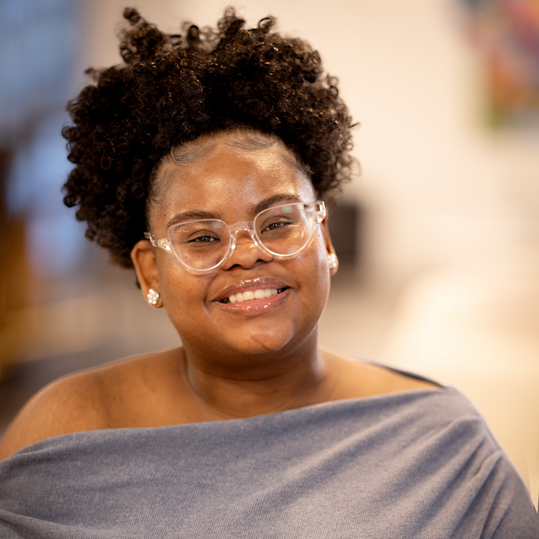 A woman with curly hair, clear glasses, and earrings smiling at the camera, wearing an off-the-shoulder gray top.