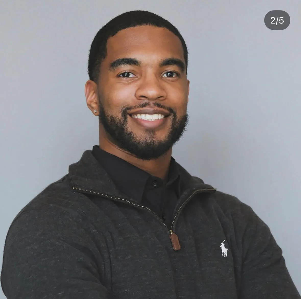 Smiling African American man with short hair and beard, wearing a dark zip-up sweater and black shirt, standing against a plain gray background.
