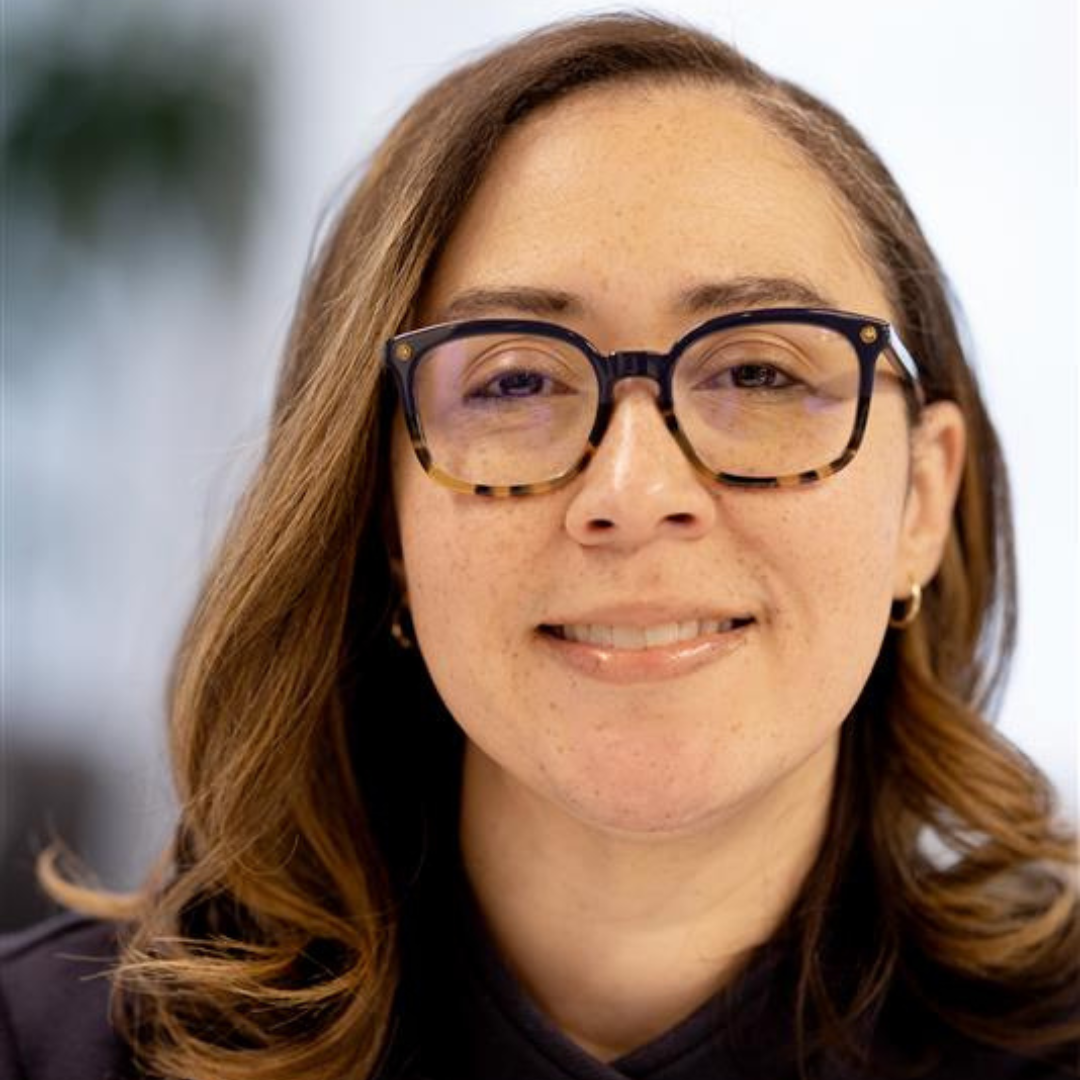 Close-up portrait of a smiling woman with light brown wavy hair wearing black-rimmed glasses with tortoiseshell accents, earrings, and a dark top, against a blurred background.