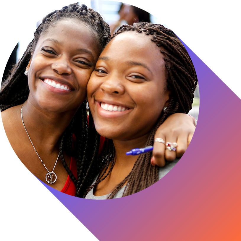 Two smiling women with braided hair close together, one with her arm around the other, in a group setting.