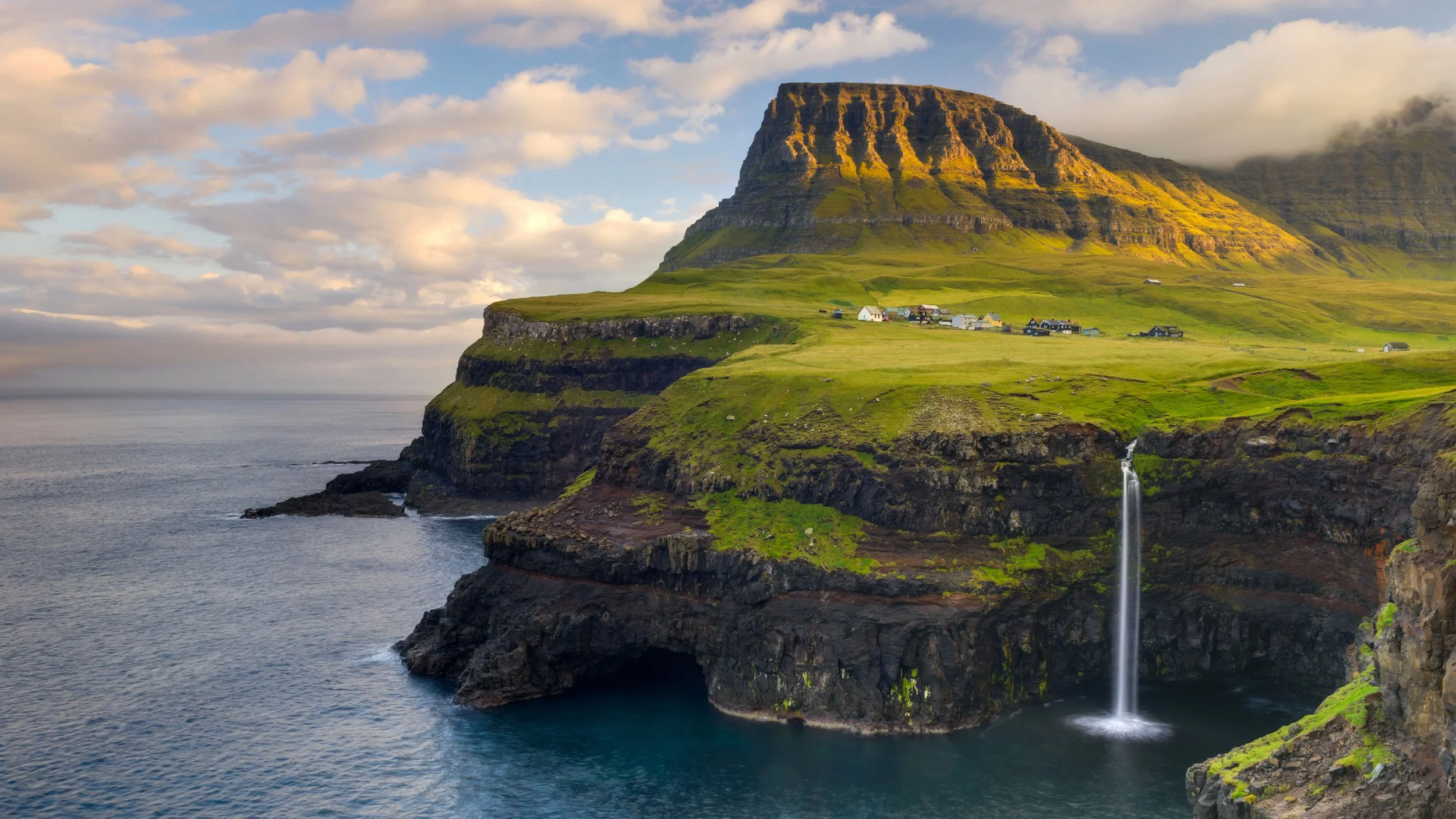 Scenic coastal landscape with green cliffs, a waterfall flowing into the ocean, and a mountain with small houses on it under a partly cloudy sky.