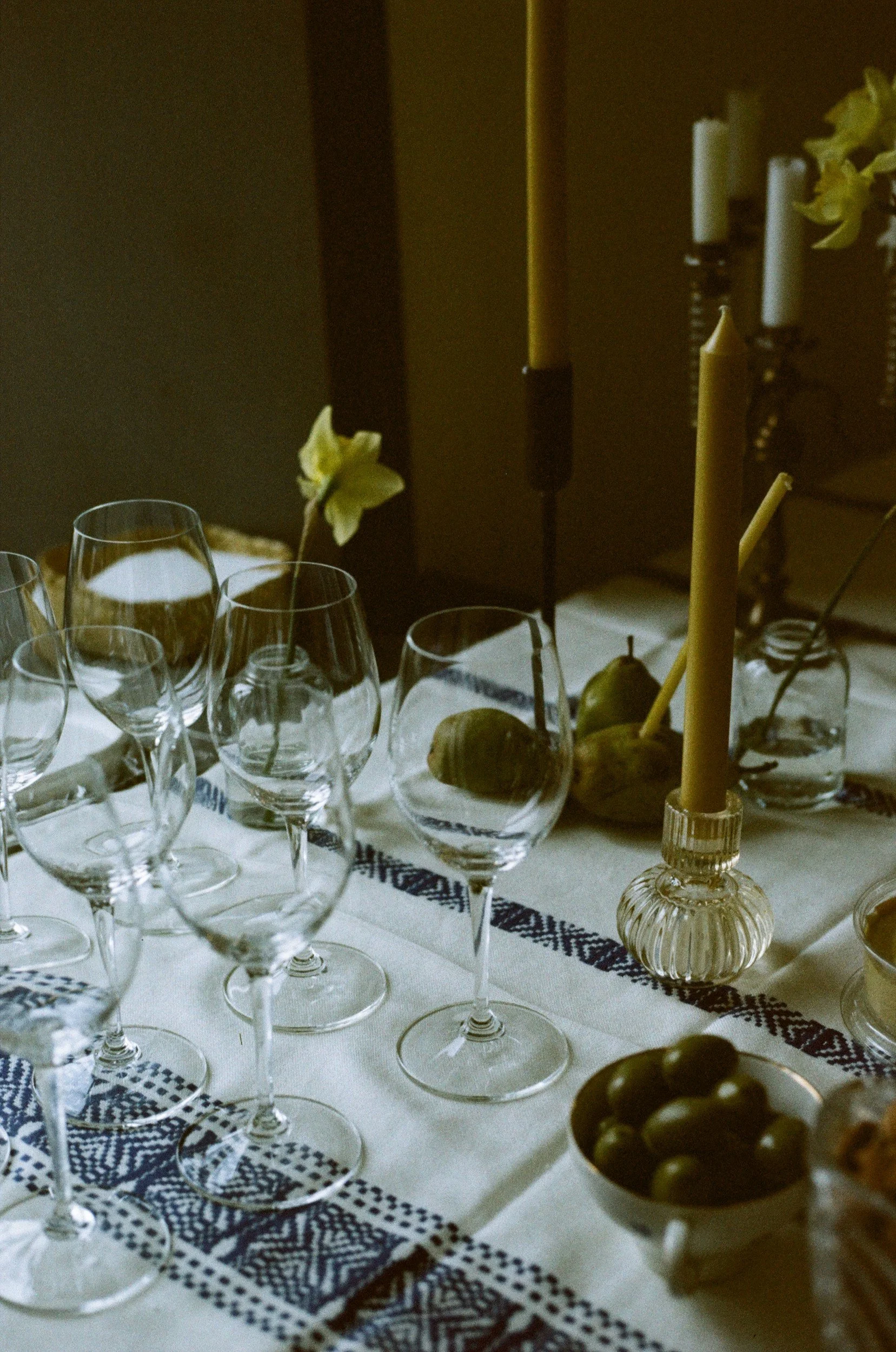 Table setting with multiple empty wine glasses, a bowl of green grapes, and yellow candles in glass candle holders, decorated with a white tablecloth and a blue embroidered table runner.