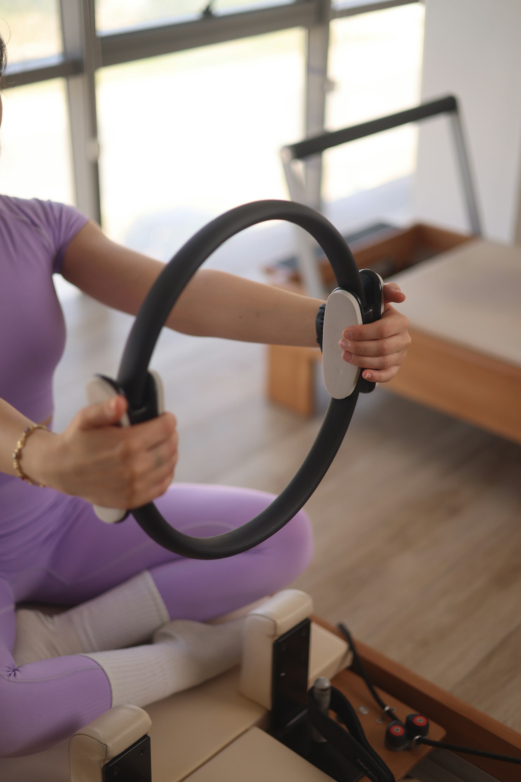 A woman in purple leggings and a lavender top using a hoop-style exercise machine in a bright room with large windows and a wooden table in the background.