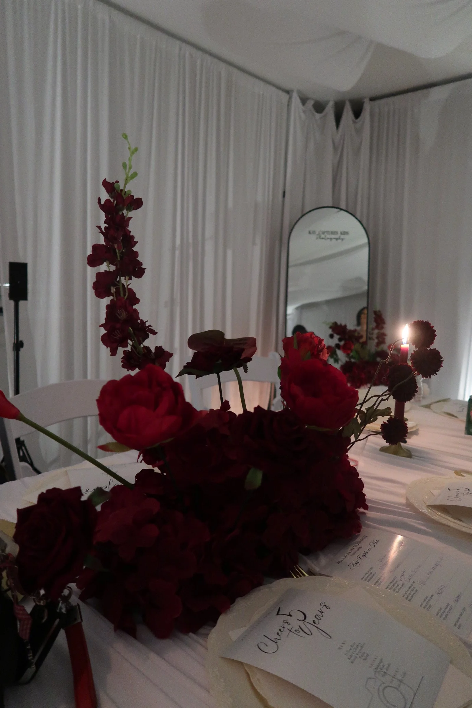 A table decorated with red floral arrangements, ceremony programs, and a white tablecloth at a wedding reception or event.