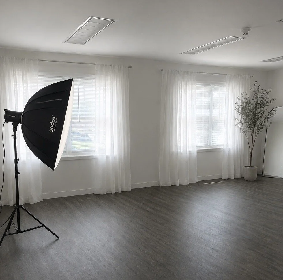 Empty room with three windows dressed in white curtains, a potted plant, a full-length mirror, and a photography studio light in the corner.