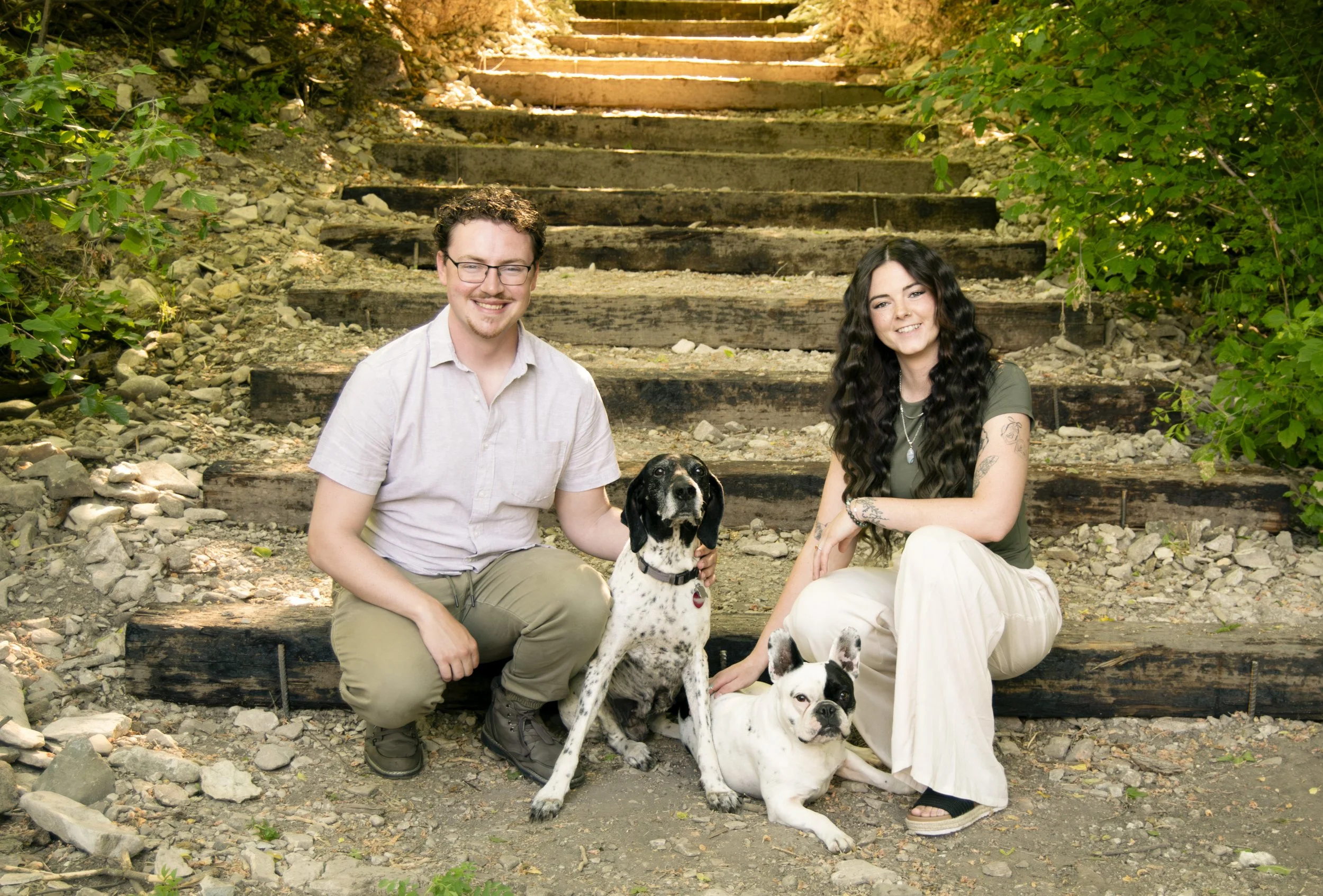 A couple, a man and a woman, sitting on wooden steps outdoors with two black and white dogs, one sitting and one lying down, surrounded by green foliage and rocky ground.