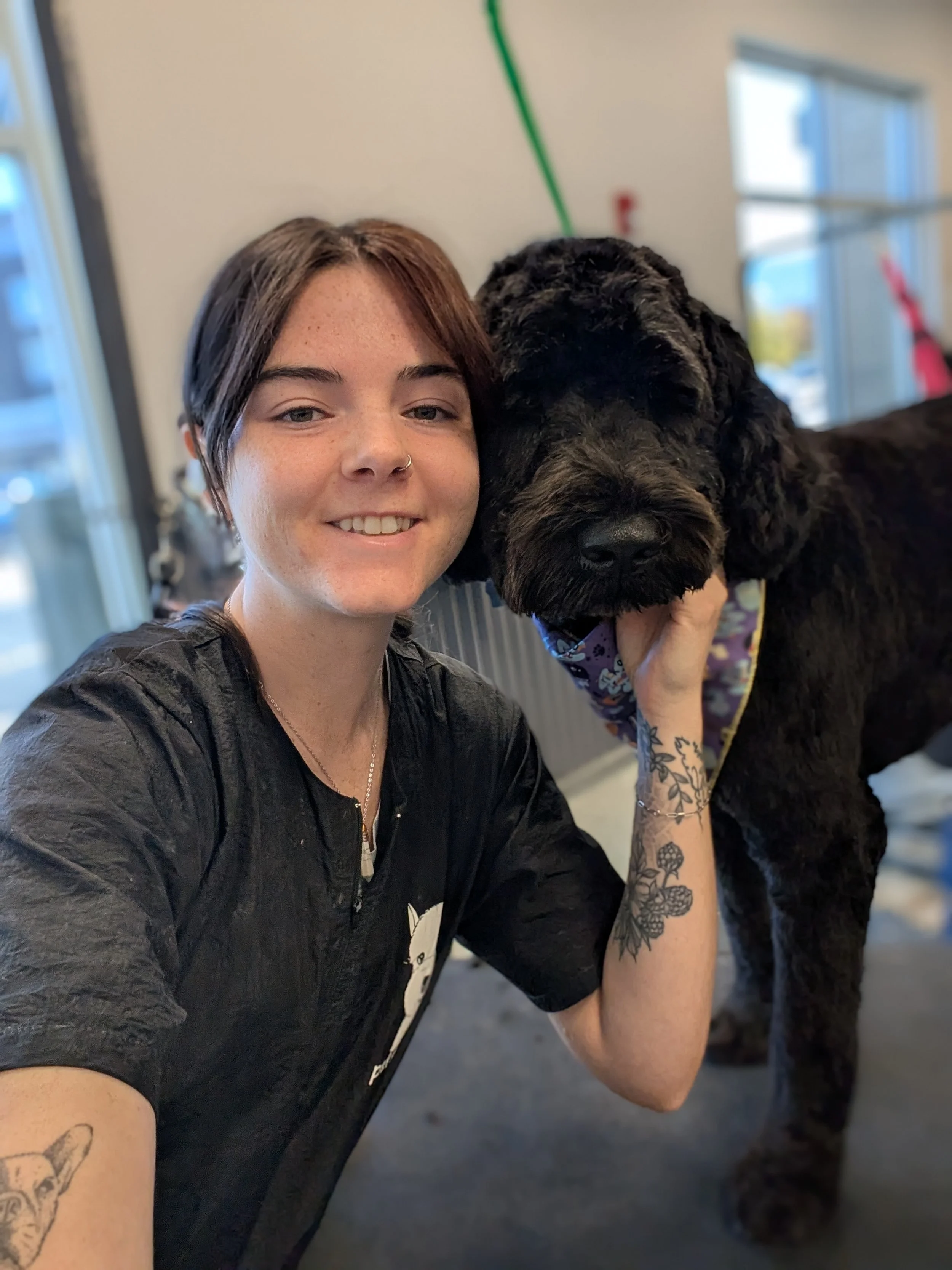 A young woman with short brown hair, smiling and taking a selfie with a large black dog that has curly fur, both in an indoor setting with windows in the background.