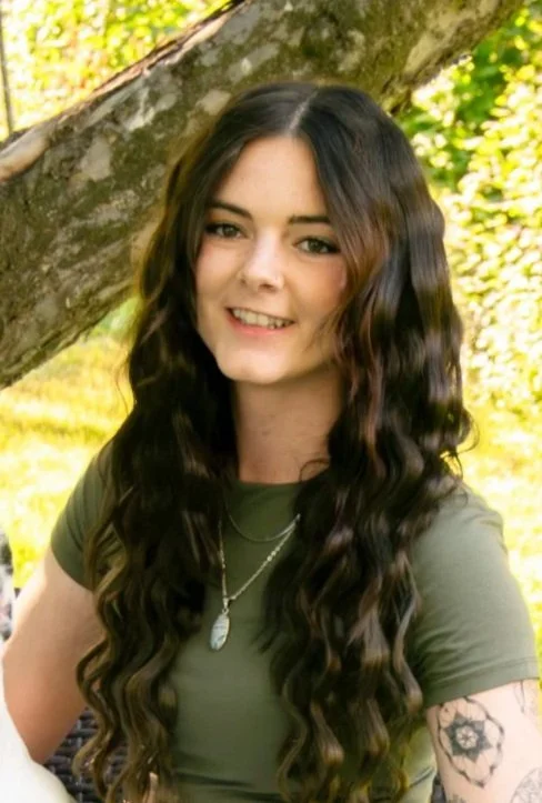 A young woman with long, dark, and curly hair, smiling outdoors near a tree with green foliage.