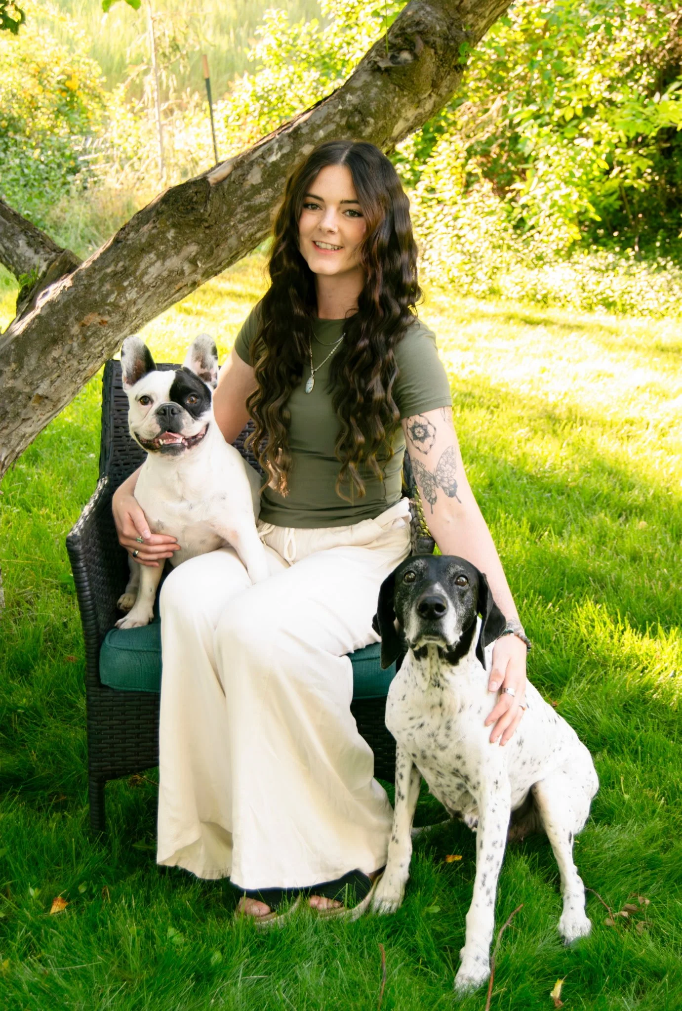 A smiling woman with long wavy brown hair, sitting on a green outdoor chair under a tree, with two dogs around her in a sunny yard with green grass and trees in the background.