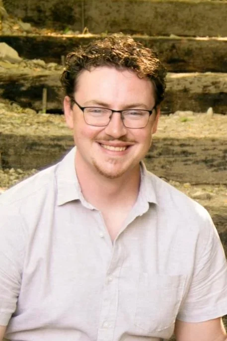 A man with curly hair, glasses, and a beard smiling outdoors, wearing a light-colored shirt with a natural background.