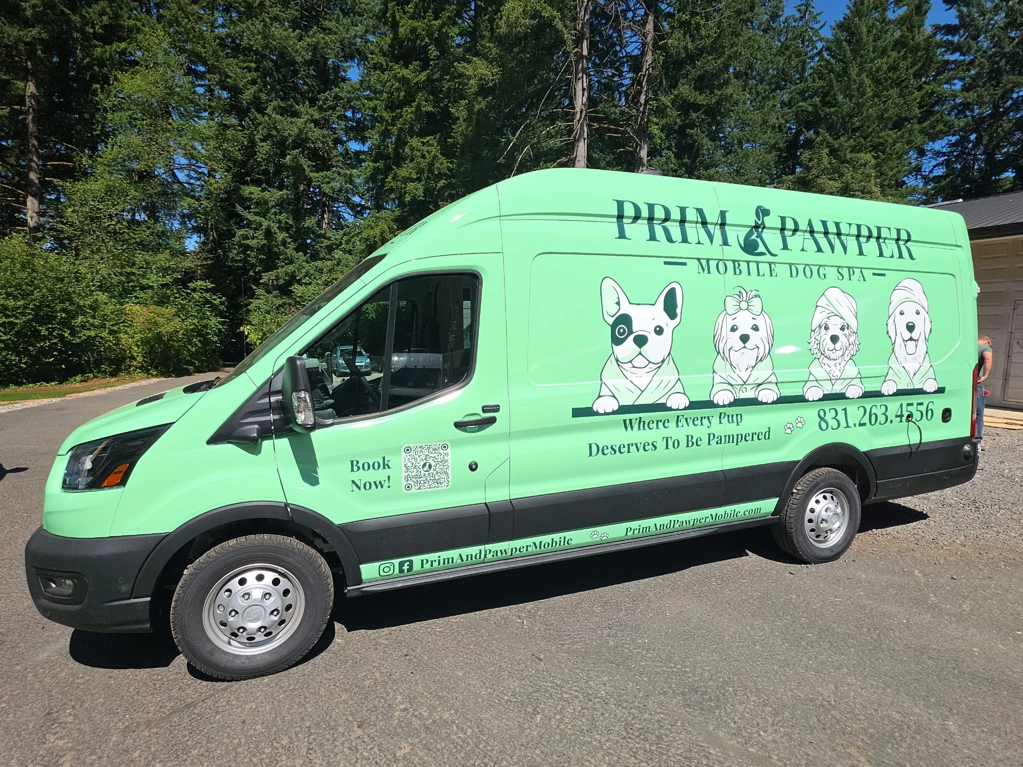 A light green mobile dog spa vehicle with cartoon images of various dog breeds on the side, parked outdoors with trees and a building in the background.