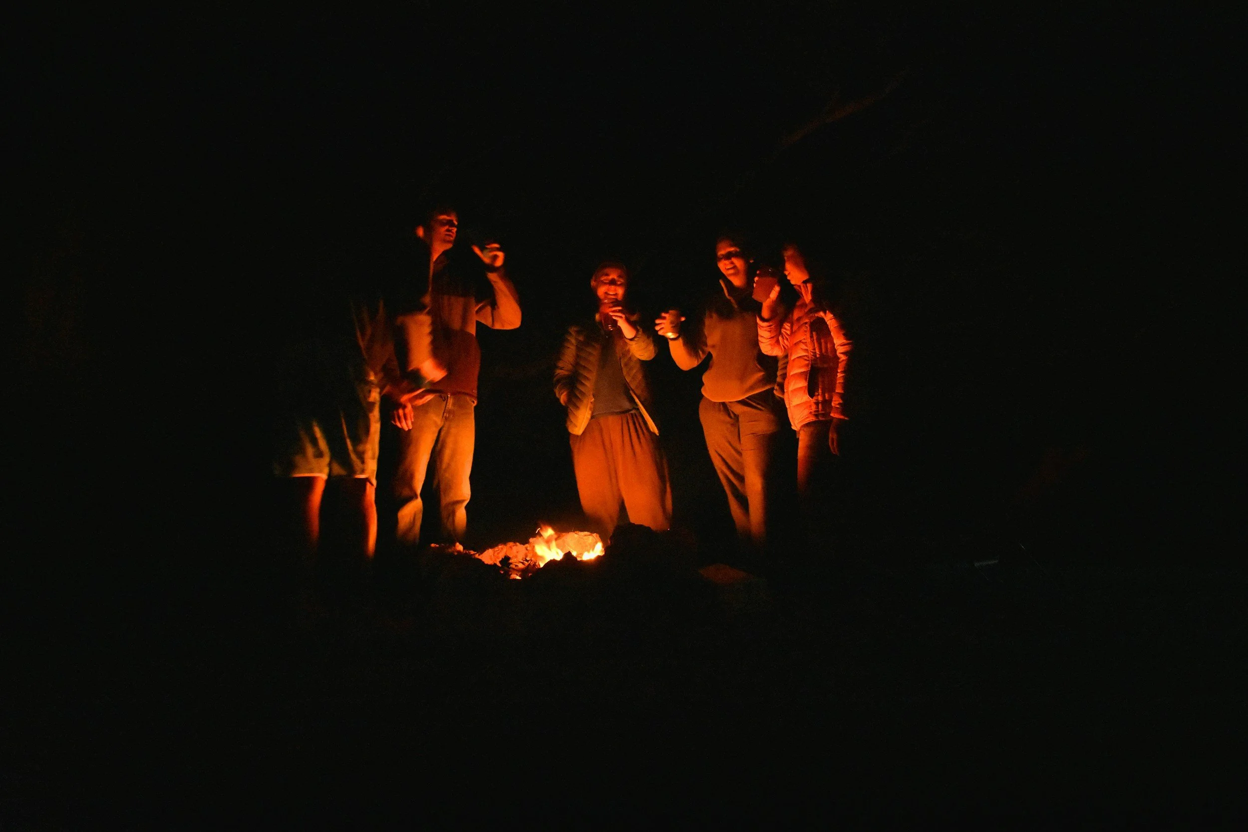 Group of people gathered around a campfire in the dark, smiling and talking.
