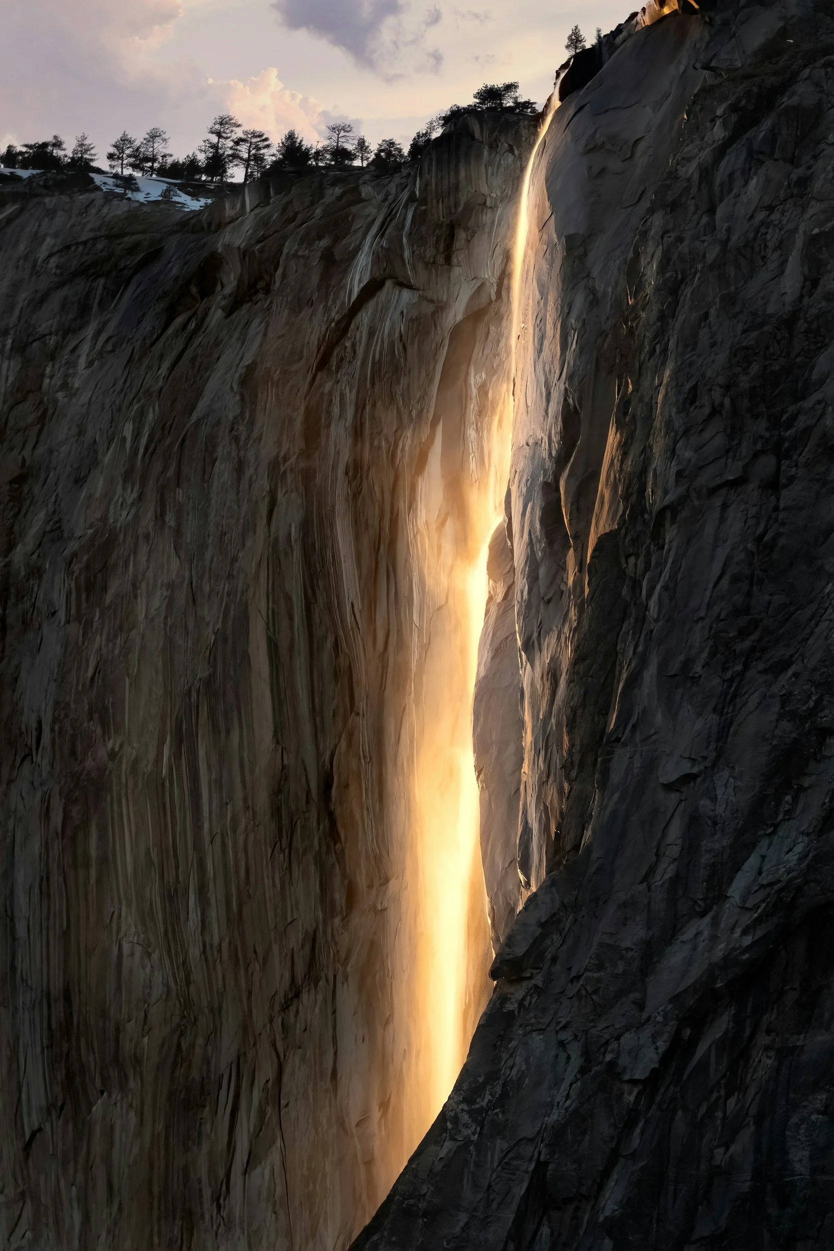 A waterfall cascading down a rocky cliff during sunset, with trees on the hilltop and clouds in the sky.