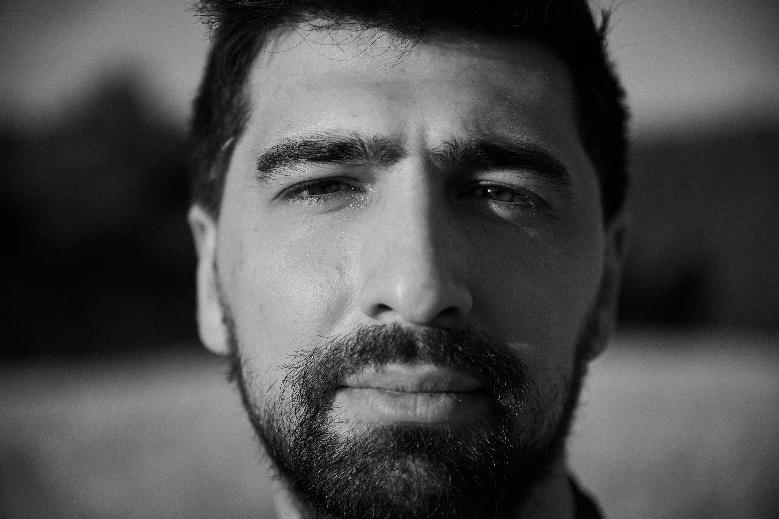Close-up of a man's face with short, dark hair and a beard, with a serious expression, in black and white.