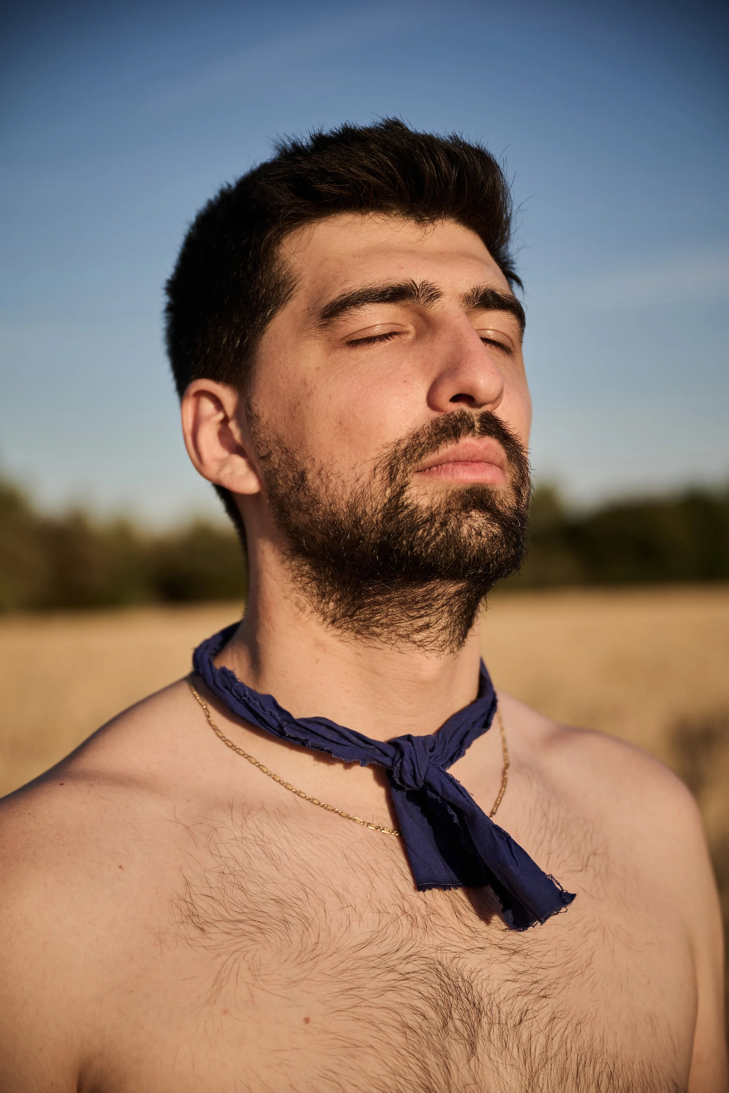 A shirtless man with a beard and closed eyes outdoors during daytime, wearing a blue cloth tied around his neck, with a blurred natural background under a clear blue sky.