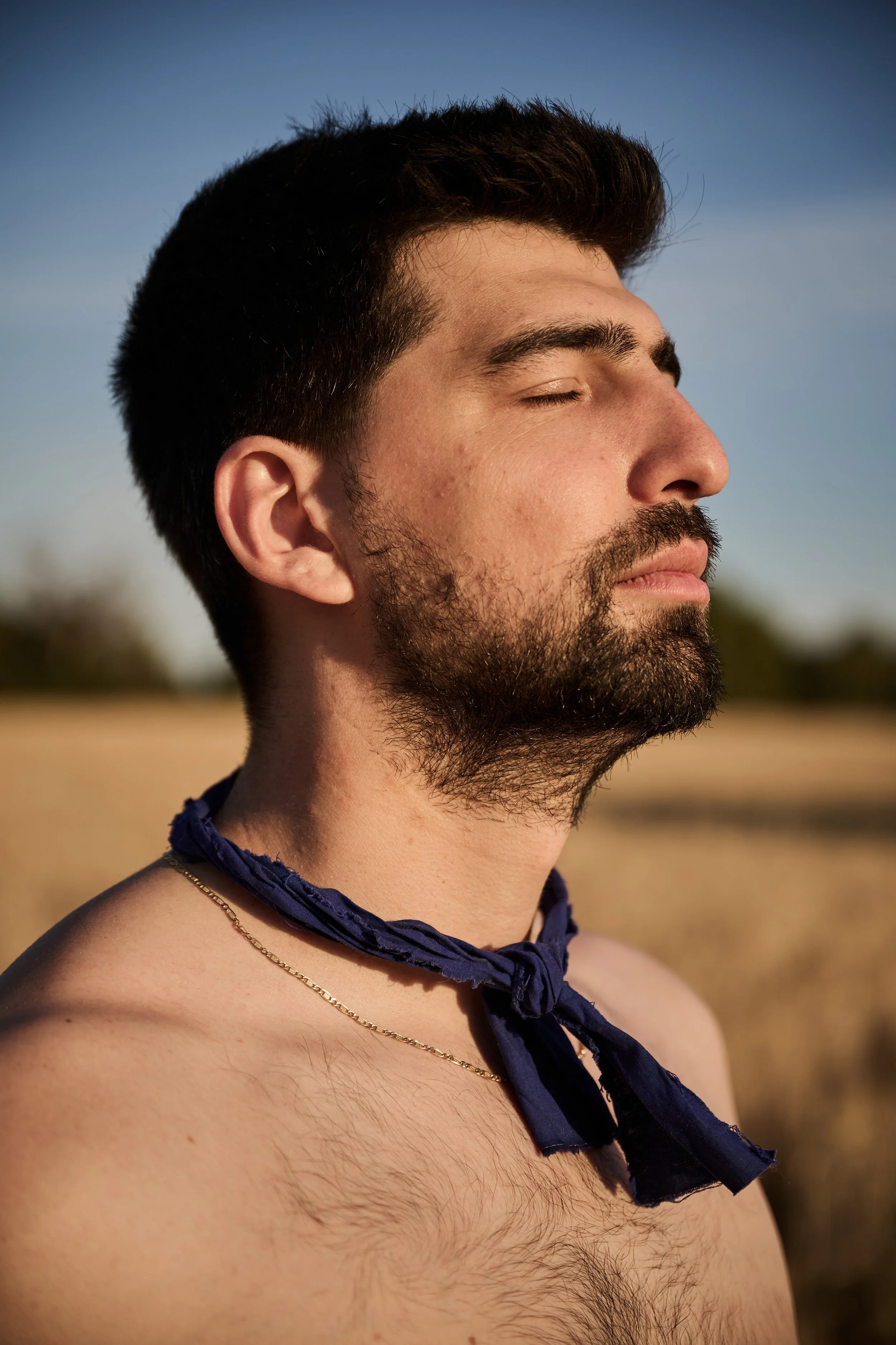 A close-up of a man with dark hair and a beard, eyes closed, outdoors with a clear blue sky in the background, wearing a blue scarf around his neck.