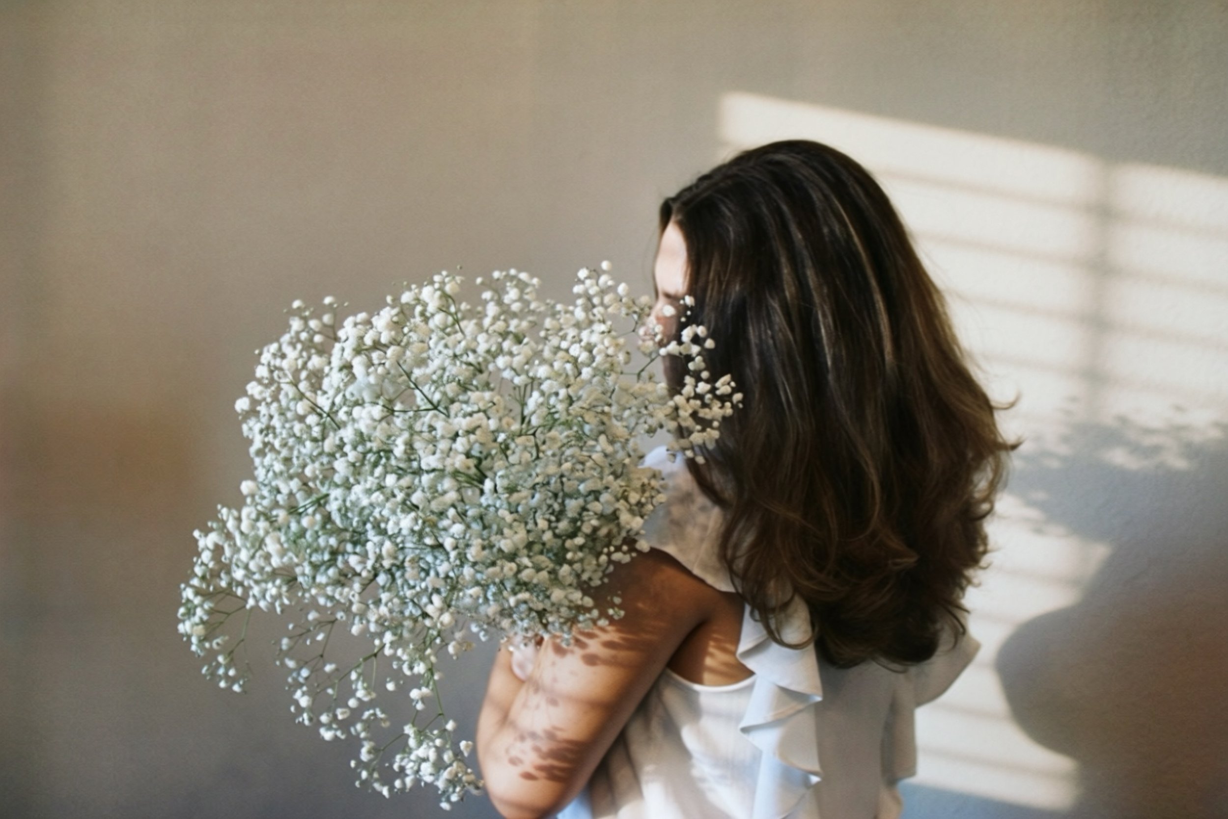 A woman with long brown hair holding a large bouquet of small white flowers, standing in front of a plain wall.