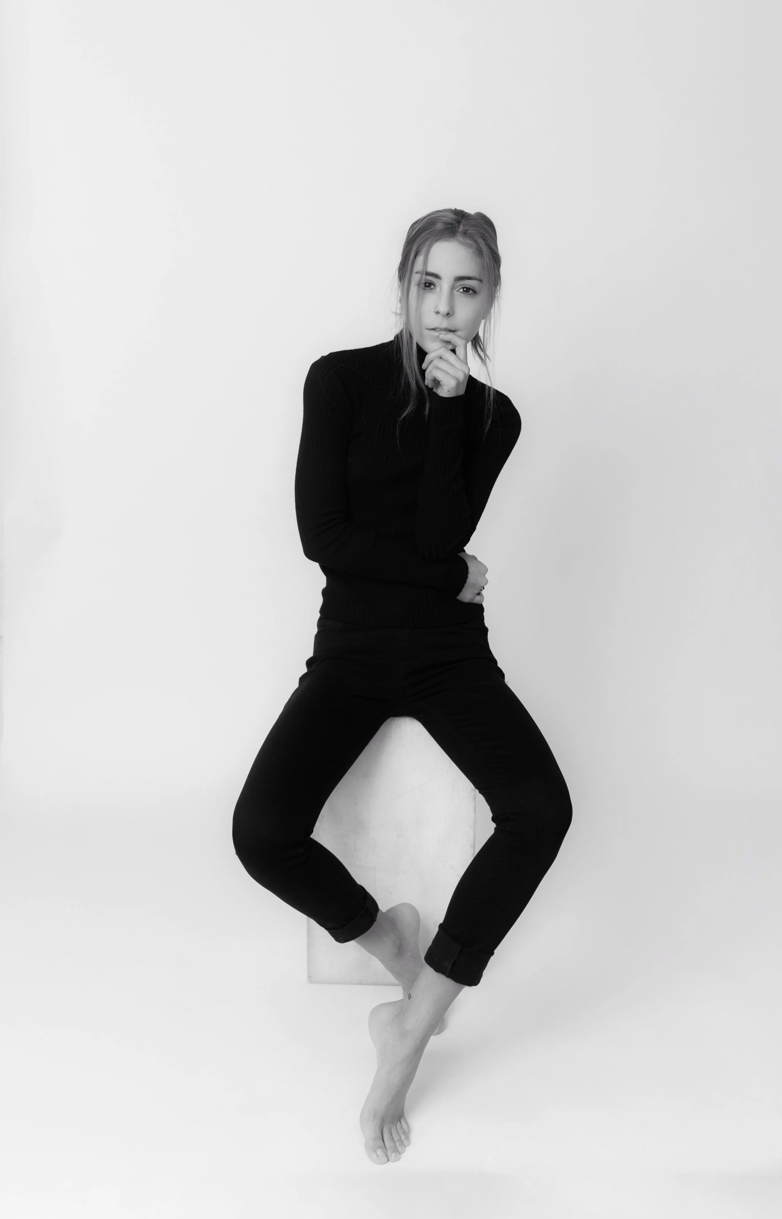 Black and white photo of a young woman with light hair, wearing a black long-sleeve top and black pants, sitting against a plain white wall with one leg bent, in a relaxed pose.