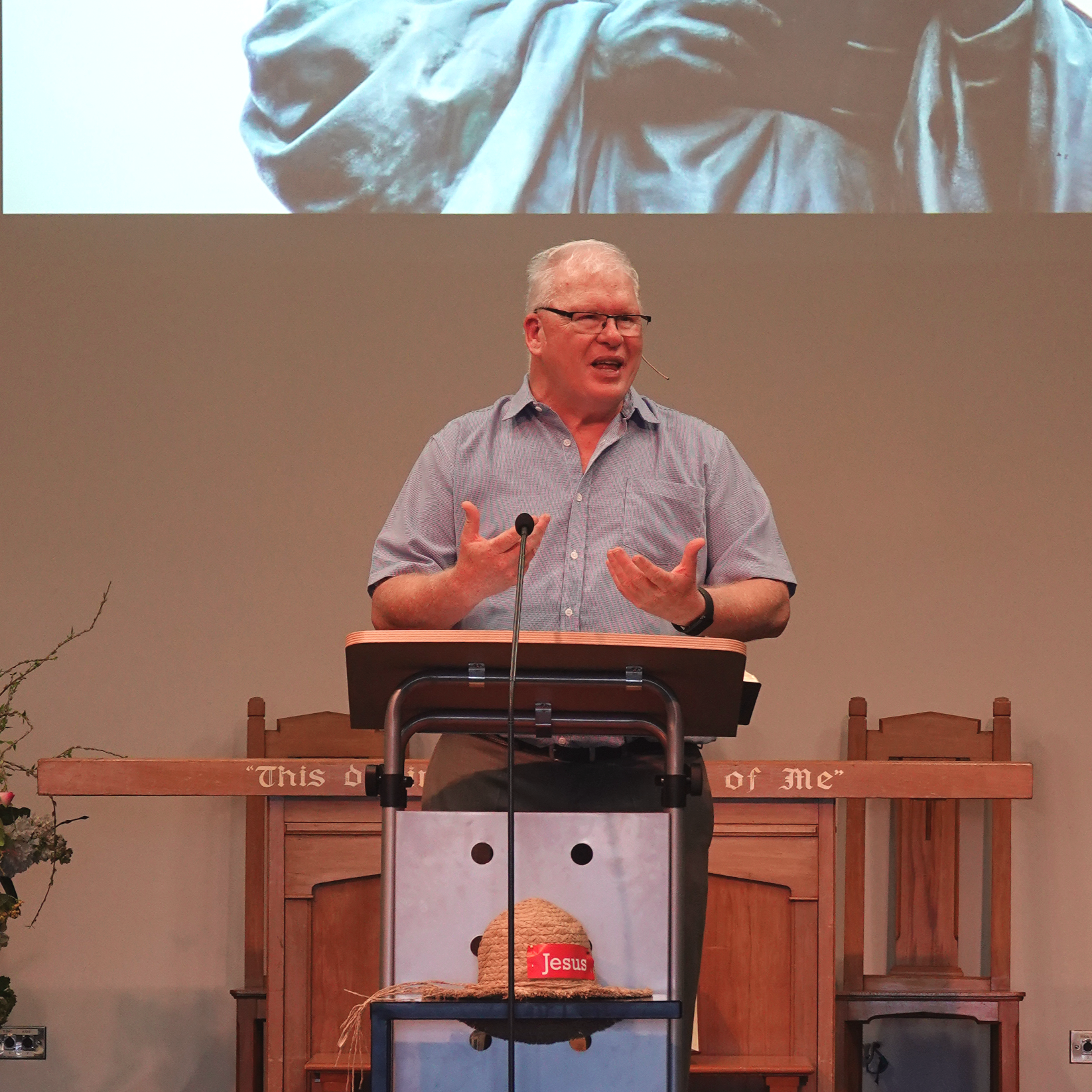 A man standing behind a pulpit giving a speech or sermon, wearing glasses and a light-colored shirt, with a church or religious setting in the background. There is a bible or religious text on the pulpit and a straw hat with a red label that says "Jesus" hanging from the front of the pulpit.