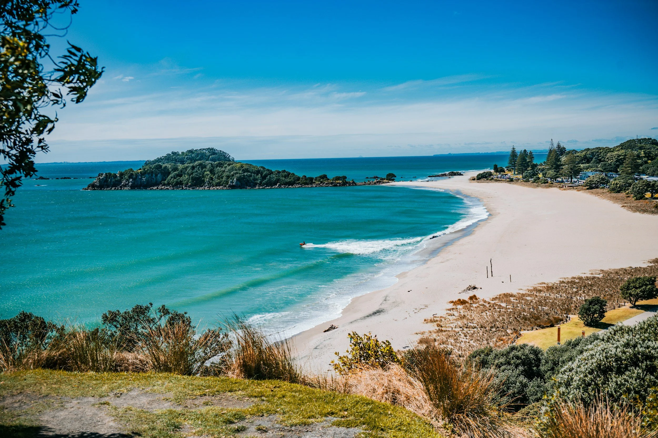 An image of taken from Mount Maunganui, New Zealand looking out towards Leisure Island. You can see the sandy beach, blue ocean and trees and grass 