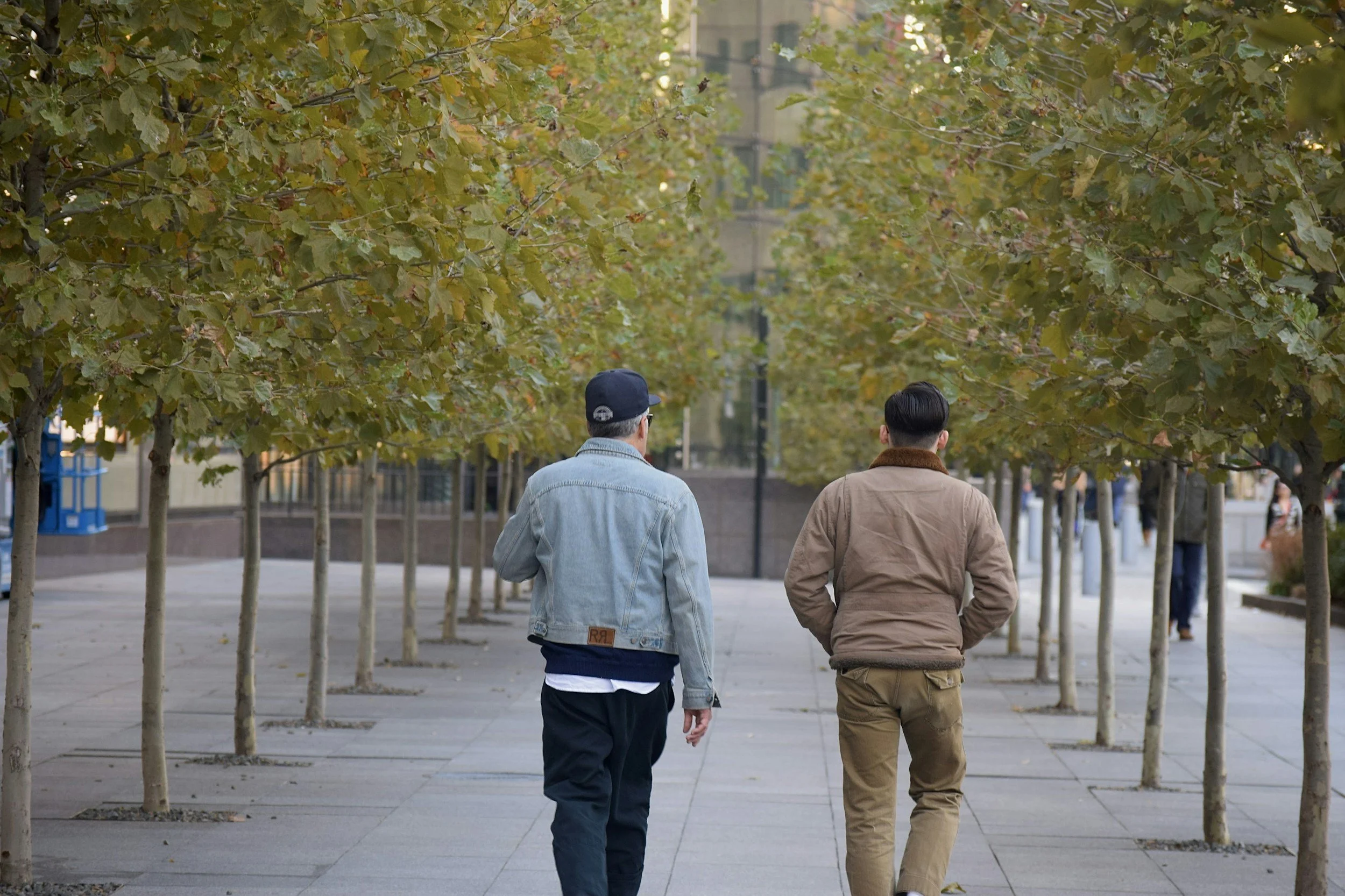 Two men walking on a sidewalk lined with trees with green and yellow leaves.