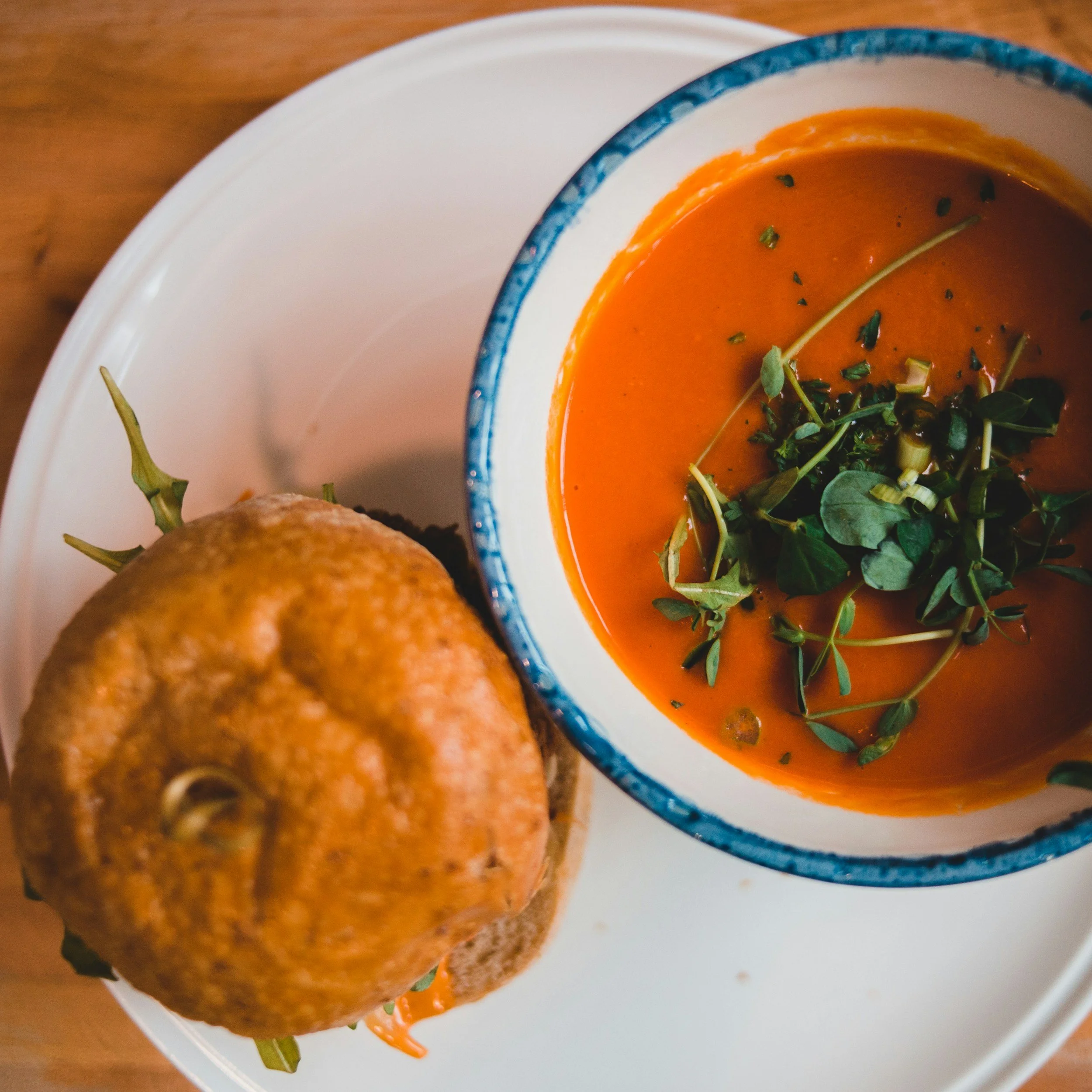 Close-up of a bowl of tomato soup garnished with herbs, with a bread roll nearby, on a white plate.