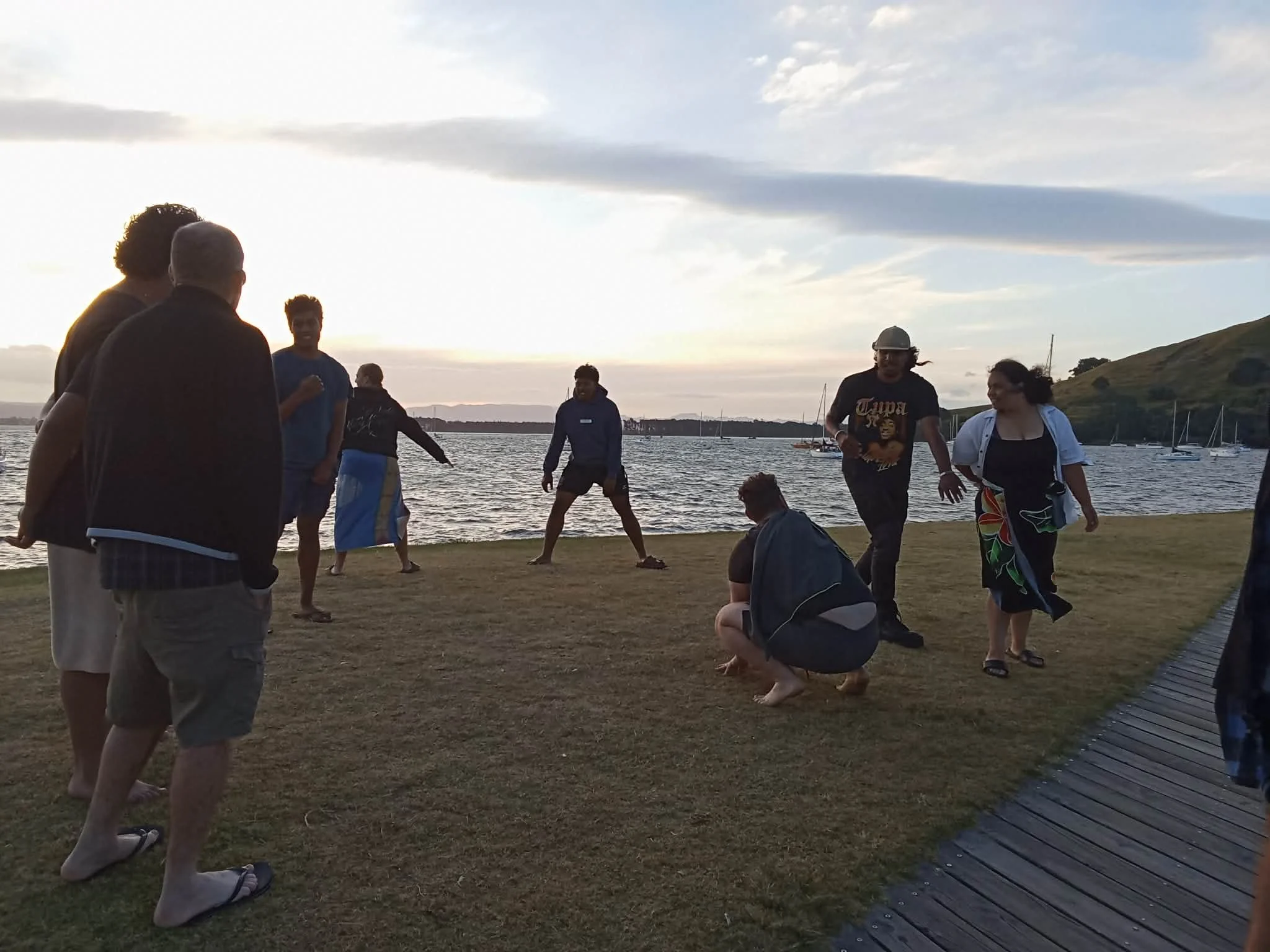 Group of people enjoying a sunset by the water, some standing, some leaning or sitting, with sailboats in the background.